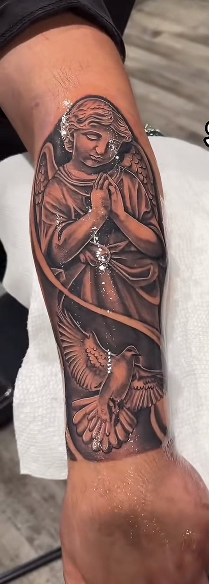 A praying angel with a dove below it, tattooed in black and grey down the inside of a forearm.