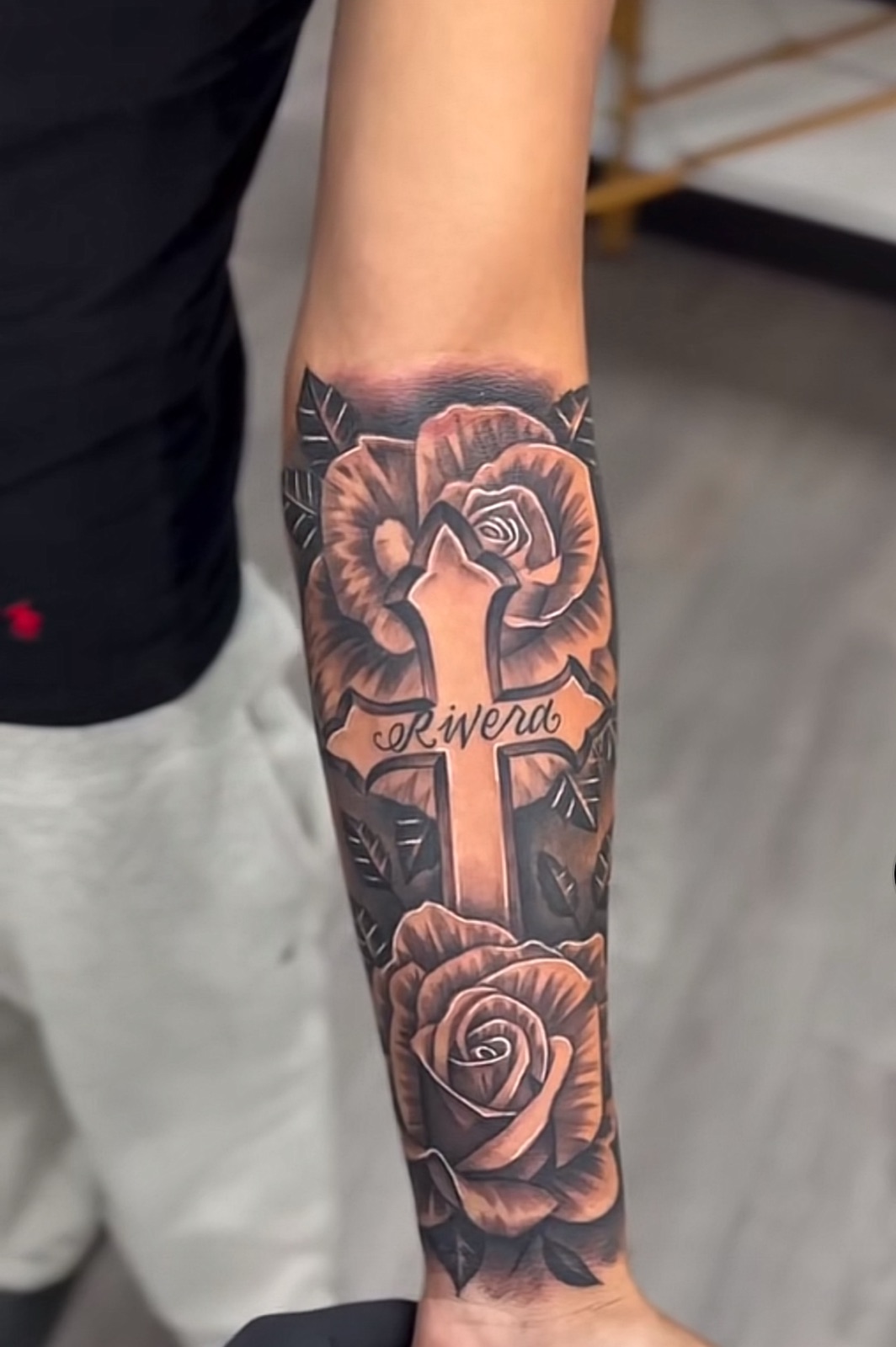 A cross reading Rivera framed by two roses, tattooed in black and grey on a forearm.